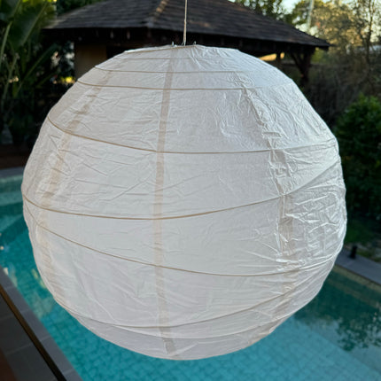 A white, irregular ribbed paper lantern hanging outdoors, displayed in an outdoor setting with a pool and trees in the background.