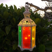 A tall hexagon LED lantern with multicolored panels hanging among greenery, illuminated from the inside with warm white lights.
